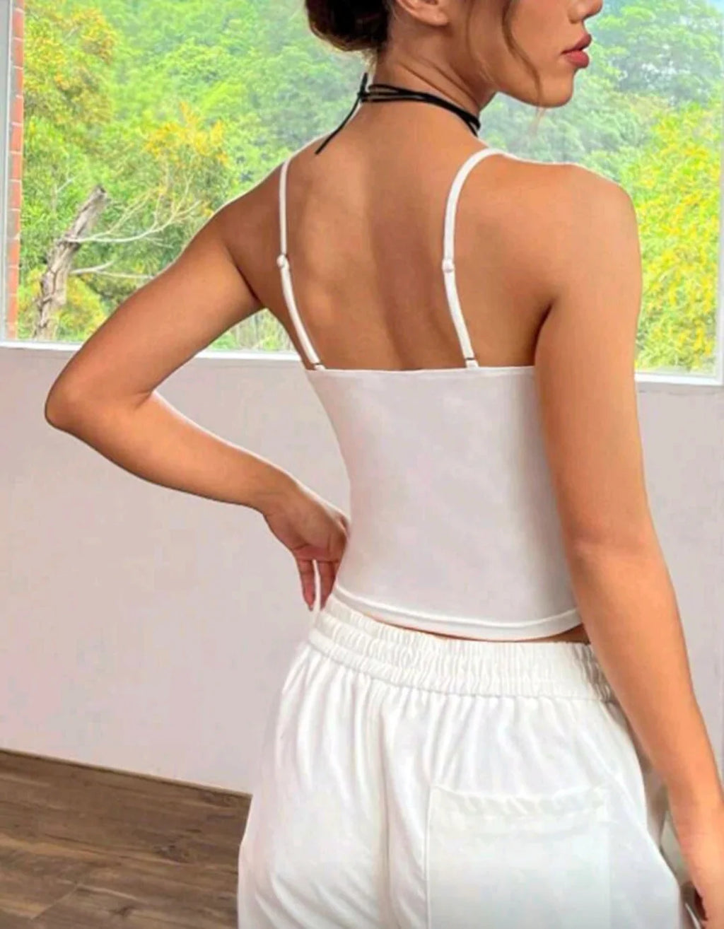 Woman in white camisole and sweatpants standing indoors with window showing green trees